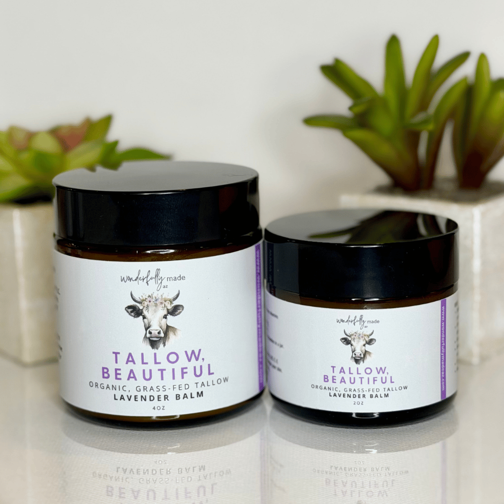 Two jars of Tallow Beautiful balm on a reflective surface with plants in the background.