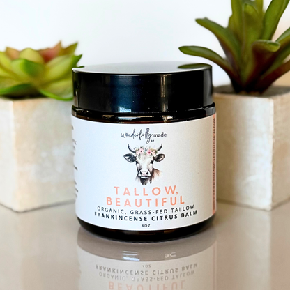 Jar of 'Tallow Beautiful' Frankincense Citrus balm with a cow illustration on a reflective surface with plants in the background.