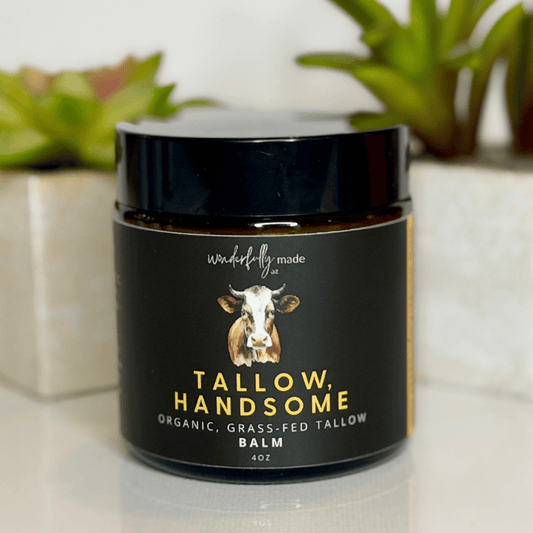 Jar of 'Tallow, Handsome' balm with a cow illustration on a white surface with plants in the background.